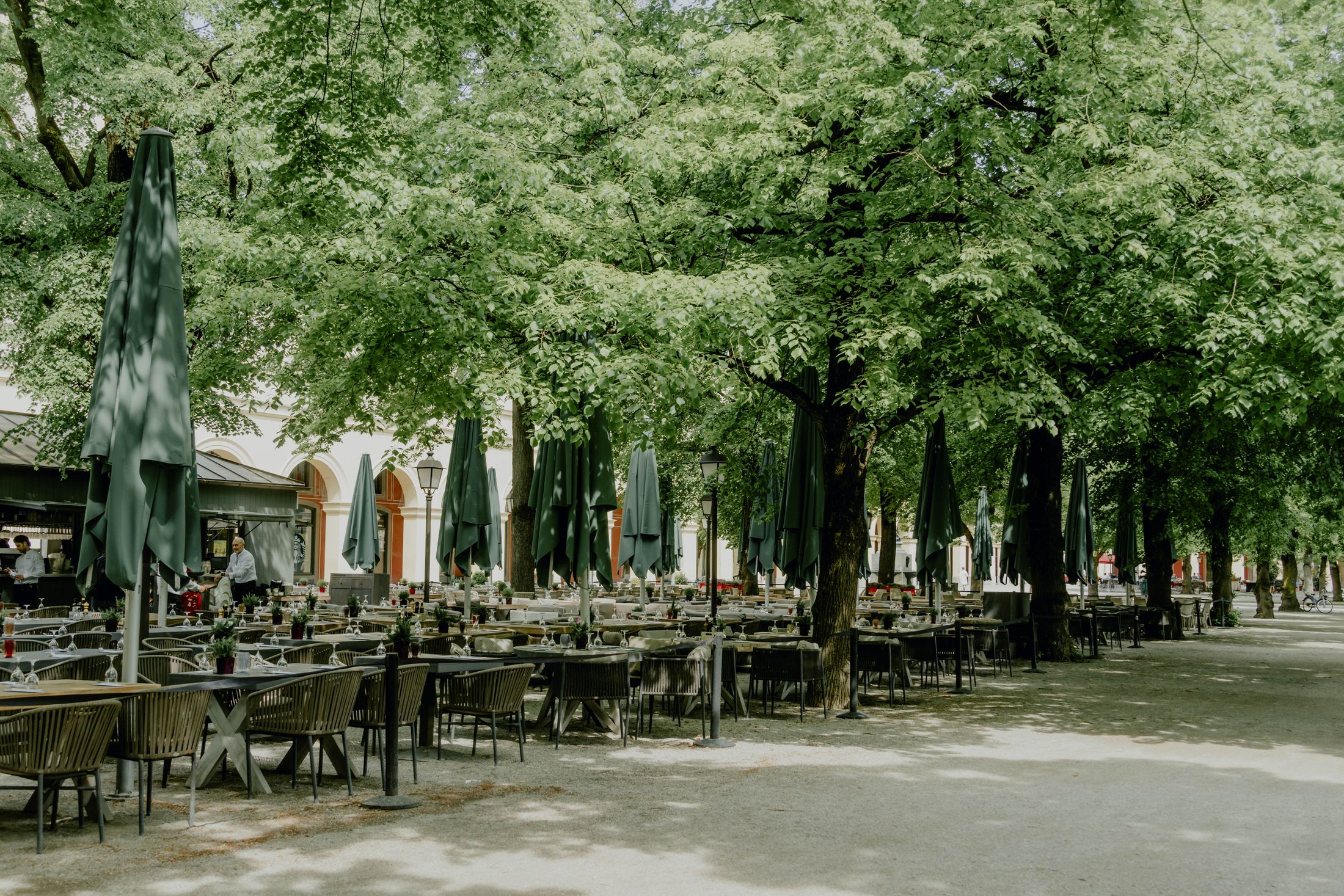 Schattiger Gastgarten mit Holztischen und Schirmen unter alten Bäumen